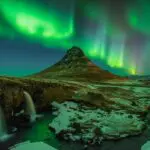 Waterfall flowing into a river with a large mountain in the background, at night with the northern lights above