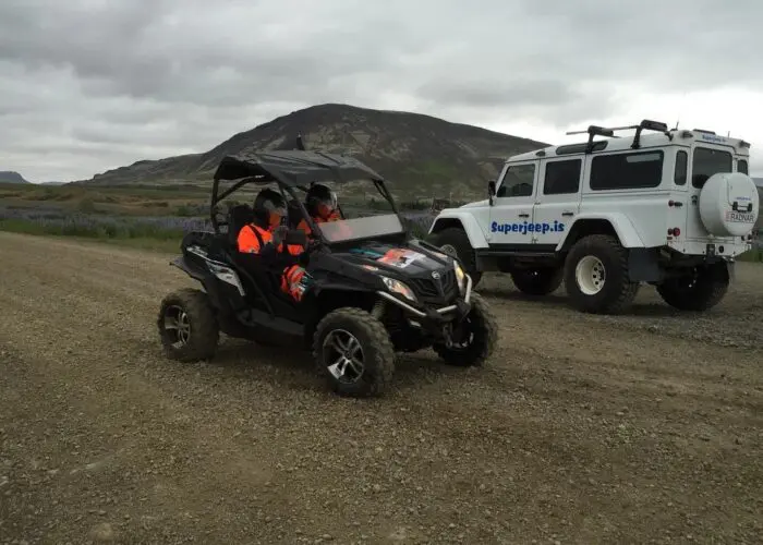 Two people in a black buggy off-roading with a superjeep parked nearby