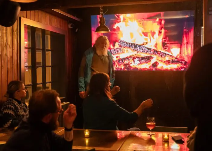 A man with long white hair giving a presentation in front of a TV displaying a cozy fireplace scene