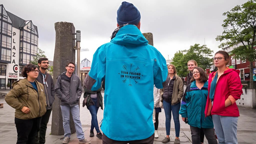 A group of tourists attentively listening to a guide wearing a blue jacket with 'Your Friend in Reykjavík' printed on the back, standing in a public square in Reykjavík. The scene includes a stone pillar and nearby buildings, including a hotel named Center Hotels Plaza. The atmosphere suggests a city tour experience in Reykjavík.