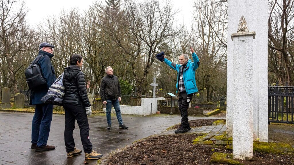 Tour guide in a blue jacket leads a group of people in a cemetery during the Reykjavik Midnight Sun Ghost Tour, with tombstones and bare trees visible in the background, capturing the haunted atmosphere of Iceland's capital.