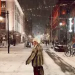 Woman turning her head to camera. While standing in the center of the road. She is wearing fur coat and jacket. In reykjavik city center. During a winter evening with snow all around.