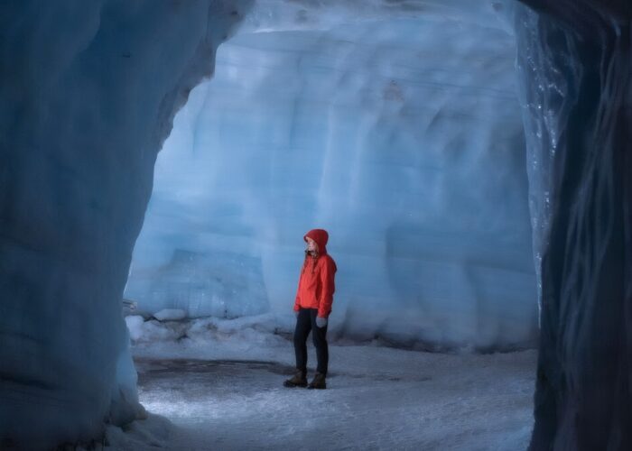 A lone visitor in a red jacket and hood standing inside a large chamber within the Langjokull glacier ice tunnel system, surrounded by tower