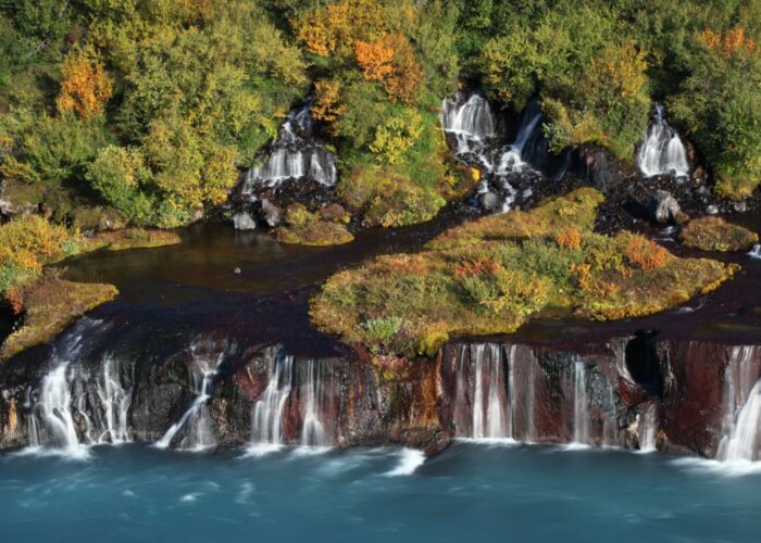 Aerial view of Hraunfossar waterfalls in autumn, with multiple cascades emerging from rust-colored and black lava rock into a vivid turquois