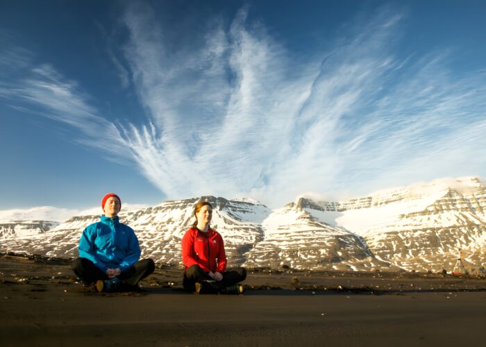 Two people sit cross-legged on dark sand in a wide mountain valley, with snow-streaked peaks and wispy clouds behind them.