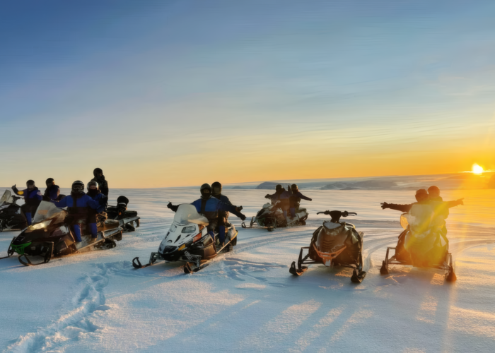 Group of snowmobile riders in blue suits paused on the vast open surface of Langjokull glacier at sunset, with a brilliant golden horizon an